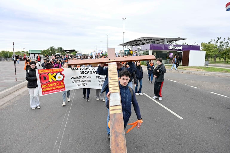 Grupo de jóvenes con ropa abrigada caminan por la calle, uno sostiene una cruz de madera y llevan un banner colorido.