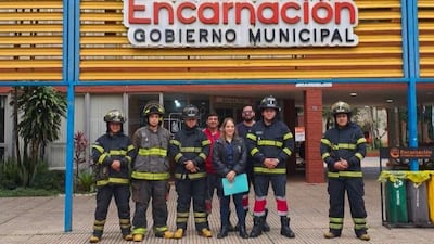Cuatro bomberos en uniforme con cascos, una mujer sostiene documentos y un hombre más, frente a Gobierno Municipal de Encarnación.