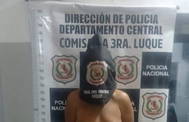 Hombre descalzo con pantalón corto de jean y cabeza cubierta con bolsa negra, frente a cartel de Policía Nacional en comisaría.