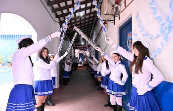Grupo de jóvenes en uniforme blanco y azul sonriendo y sosteniendo cintas coloridas en un pasillo escolar.