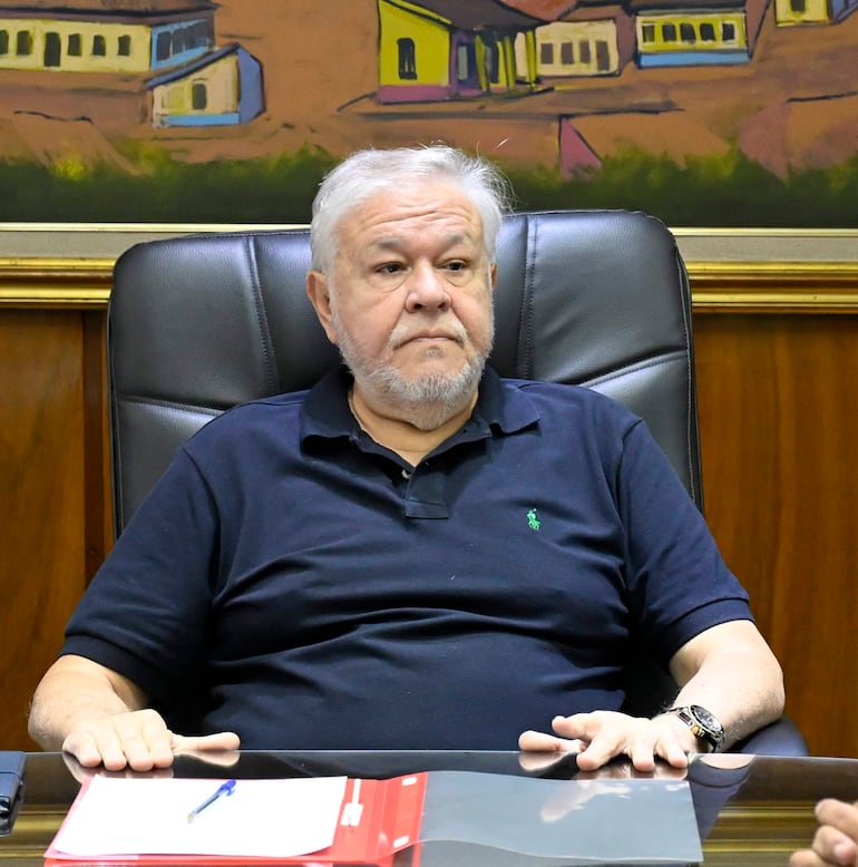 *Jorge Brítez, presidente de IPS Hombre canoso con barba, camiseta negra y pantalón claro, sentado serio en sillón de oficina con documento y bolígrafo frente a él.