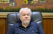 *Jorge Brítez, presidente de IPS Hombre canoso con barba, camiseta negra y pantalón claro, sentado serio en sillón de oficina con documento y bolígrafo frente a él.