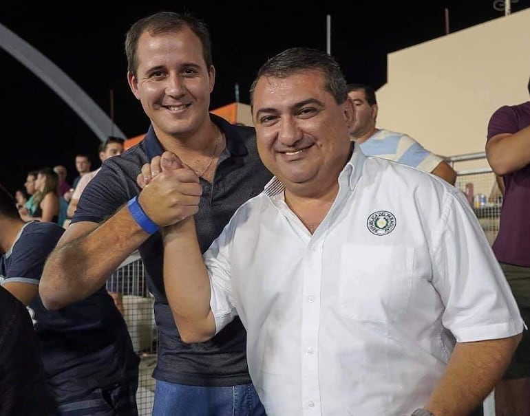 Carlos Alberto Pereira Rieve sonriente en camisa oscura y Luis Yd en camisa blanca con logo, dándose la mano en un evento nocturno.
