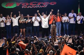 Ocho hombres aplauden en el escenario, con uno en el centro levantando los brazos. La audiencia sostiene banderas naranjas.