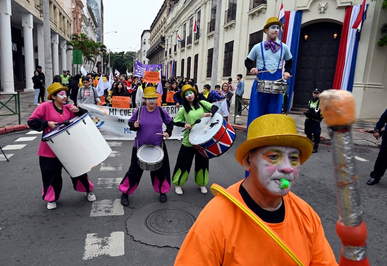 Tres payasos coloridos, uno con bombo blanco y otro con tambor rojo, animan la protesta en un ambiente festivo en Asunción.