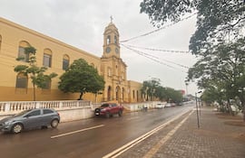 Edificio amarillo de iglesia con torre y reloj, rodeado de árboles y vehículos estacionados, en un día lluvioso.