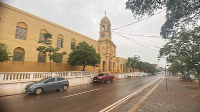 Edificio amarillo de iglesia con torre y reloj, rodeado de árboles y vehículos estacionados, en un día lluvioso.