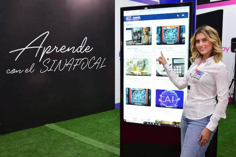 Mujer rubia y ondulada en camisa blanca y jeans, señalando pantalla con contenido educativo en ambiente moderno.