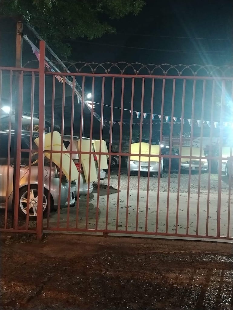 Reja metálica roja cierra entrada a estacionamiento oscuro, con varios vehículos cubiertos con cartón en su parte trasera.