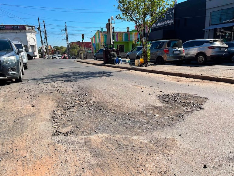 Calle urbana con un bache grande, vehículos grises y azules estacionados, edificios coloridos de fondo y cielo despejado.