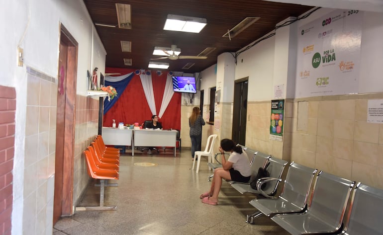 Mujer sentada pensativa en silla blanca, otra mujer de pie hablando en un espacio iluminado con decoración roja y azul.