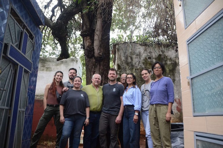 Nueve personas de pie junto a un árbol, algunas con ropa de vaqueros, lucen serias y esperanzadas en un ambiente informal.