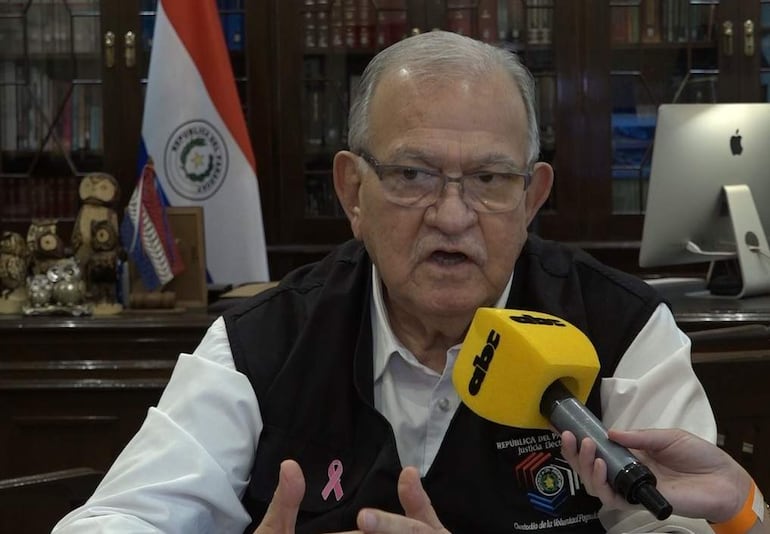 Jorge Bogarín González con chaleco oscuro y gafas, hablando intensamente frente a un micrófono en un entorno institucional decorado.