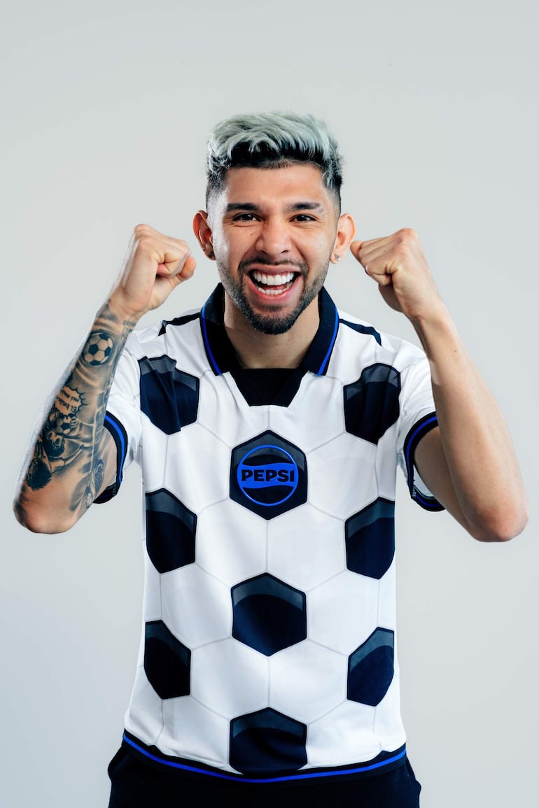 Julio Enciso sonríe mientras viste camiseta blanca con diseño hexagonal de Pepsi, levantando los brazos en un fondo neutro.