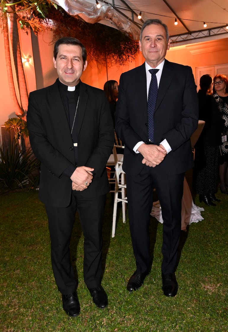 Sacerdote de sotana y hombre con traje oscuro en evento festivo al aire libre, rodeados de decoración elegante y luces.