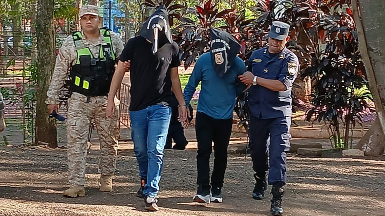 Dos hombres con capuchas, uno en camiseta negra y otro en camiseta azul, caminando juntos, escoltados por un policía y un guardia.