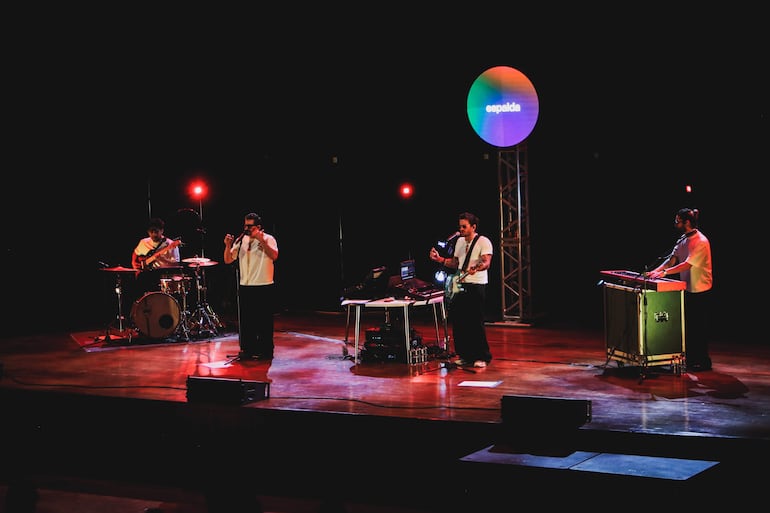 Cuatro músicos en escenario oscuro, iluminado por luces de colores. Vocalista con micrófono y guitarrista en camisetas blancas.