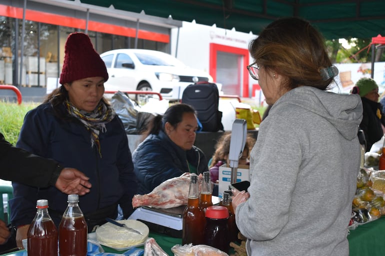 Mujer con chaqueta oscura y bufanda compra en un puesto, mientras otro hombre le muestra productos sobre la mesa.