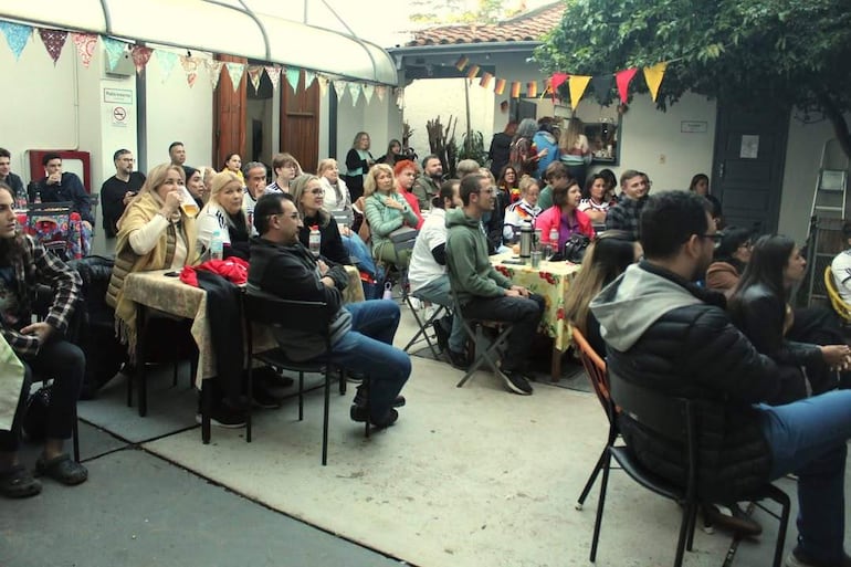 Aproximadamente 30 personas sentadas en un ambiente festivo, algunas conversando y otras mirando hacia un área de presentación.