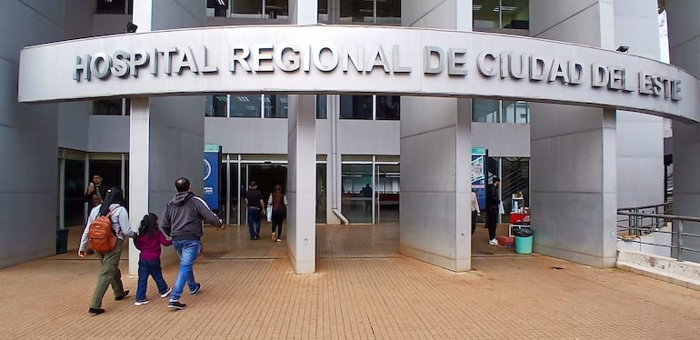 Adulto con mochila y niño de la mano, caminando frente al moderno edificio del Hospital Regional de Ciudad del Este.