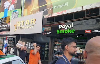 Hombre con camiseta naranja lleva cajas, mientras otro hombre vestido de negro interactúa en una zona comercial céntrica.