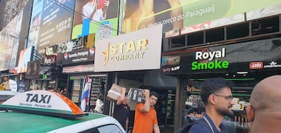 Hombre con camiseta naranja lleva cajas, mientras otro hombre vestido de negro interactúa en una zona comercial céntrica.