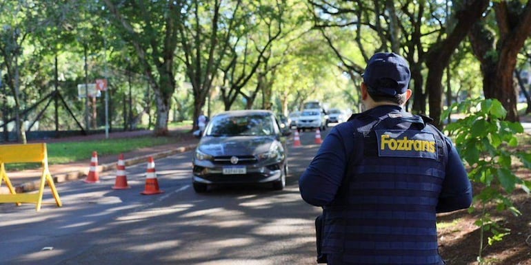 Hombre de espaldas en chaleco Foztrans dirigiendo el tráfico, rodeado de vehículos y árboles en un ambiente tranquilo.