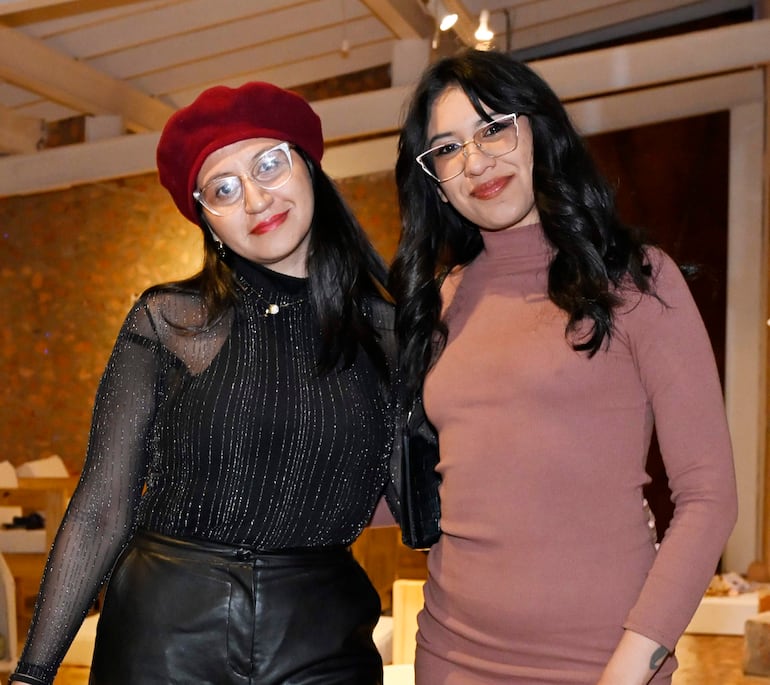 Dos mujeres, una con sombrero rojo y blusa negra, otra en vestido rosa, sonríen en un entorno moderno y festivo.