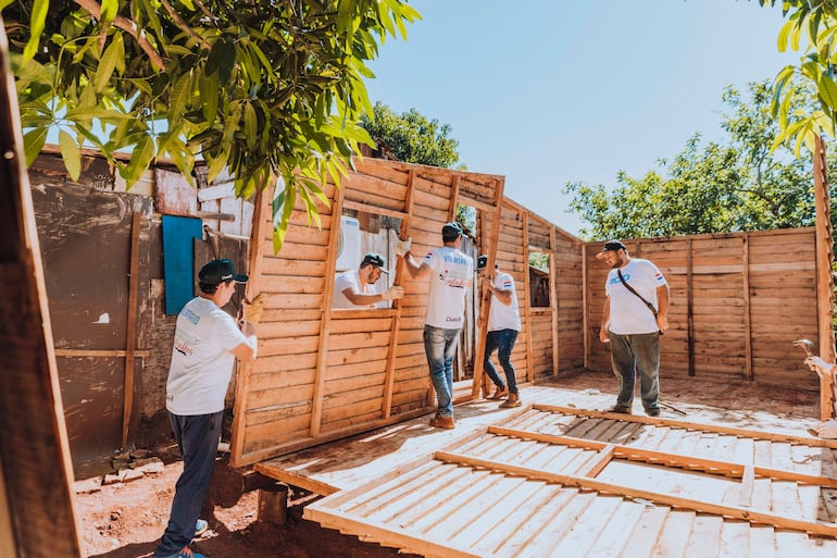 Cinco voluntarios en camisetas blancas colaboran en la construcción de una estructura de madera bajo un sol brillante y vegetación alrededor.