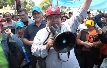Gabriel Espínola con megáfono y gorra roja, rodeado de personas con pancartas en una manifestación exterior.