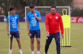 Antolín Alcaraz gesticula en camiseta roja, mientras dos jóvenes con camisetas azules lo escuchan atentos en el campo.