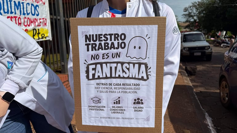 Dos bioquímicos con batas blancas sosteniendo un cartel en la calle, en medio de una manifestación urbana.