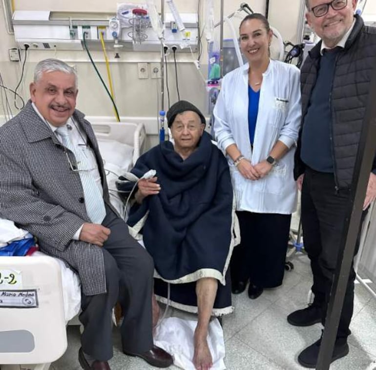 Monseñor Melanio Medina sentado en cama, rodeado de sonrientes visitantes, en un ambiente de hospital.