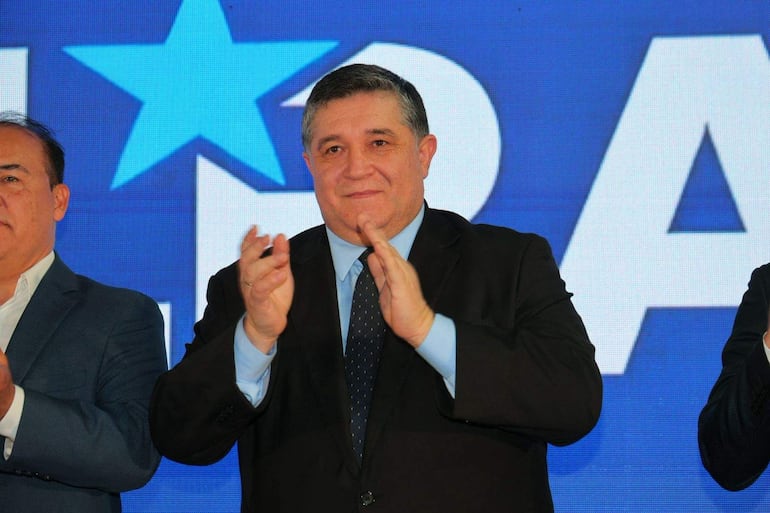 Alcides Riveros en traje oscuro, aplaudiendo con expresión seria junto a dos hombres en traje y chaqueta, ante fondo azul y blanco.