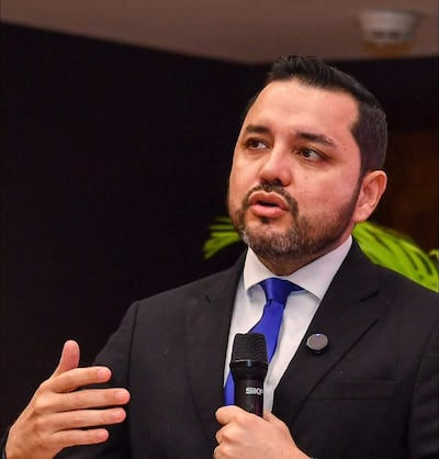 Luis Bello en traje oscuro y corbata azul, con gesto enfático mientras sostiene un micrófono en un salón de conferencias.