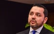 Luis Bello en traje oscuro y corbata azul, con gesto enfático mientras sostiene un micrófono en un salón de conferencias.
