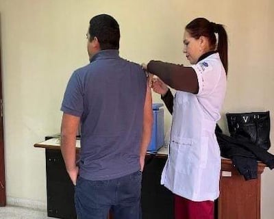 Hombre de espaldas con camiseta azul oscuro recibe vacuna de mujer en bata blanca, ambiente interno de salud, centrados en la tarea.