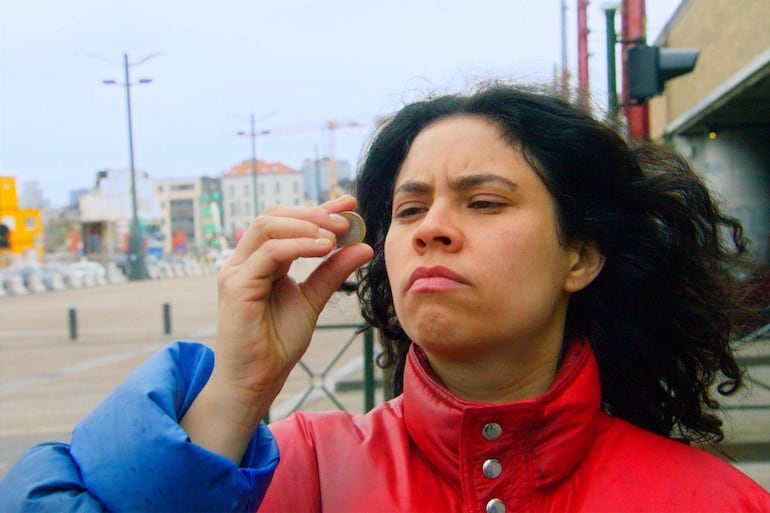 Mujer con abrigo rojo examina una moneda en un área urbana bajo un cielo nublado, sin otras personas alrededor.