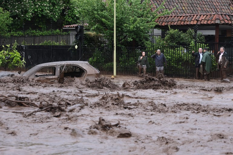 Personas observan un vehículo afectado por un aluvión este jueves, en la comuna de Las Condes, en Santiago (Chile). Las autoridades chilenas informaron que, en medio de las fuertes lluvias que afectan la zona centro-sur del país, se produjo un aluvión en una zona precordillerana al este de Santiago, que arrastró vehículos y buses. EFE/ Javier Torres