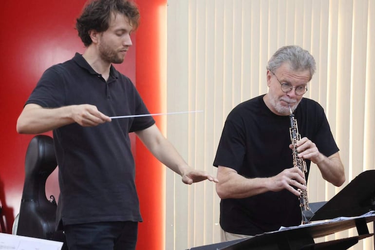 Hombre mayor con gafas y barba toca oboe d'amore, mientras un joven en camiseta negra dirige con batuta.