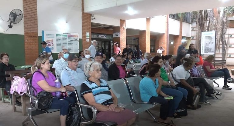 Grupo de adultos mayores, algunos con mascarillas, esperan en bancos de metal en un ambiente tranquilo del hospital.