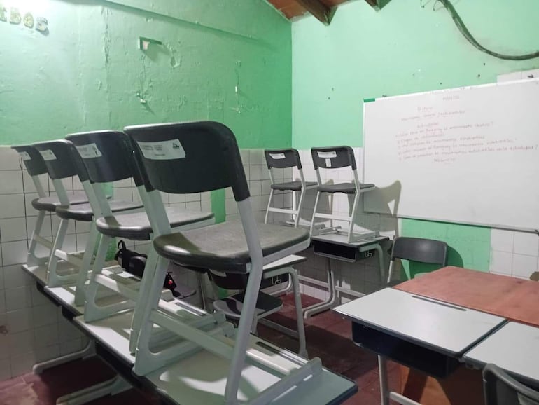 El baño reconvertido en aula. La misma está vacía con sillas apiladas sobre mesas y pizarra blanca. En las paredes se ven aún los azulejos del sanitario desmantelado.