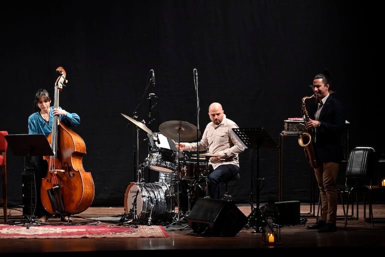 Músicos en el escenario: mujer con contrabajo, hombre calvo en batería y otro con saxofón, todos en un ambiente iluminado.