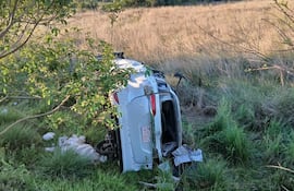 Vehículo volcado en un área rural, rodeado de pasto alto y vegetación. No hay personas visibles.