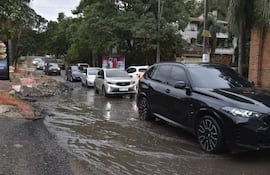 Seis autos atascados en agua, calle deteriorada con escombros y área de construcción a la izquierda, día nublado.