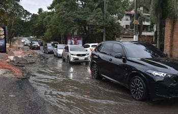 Seis autos atascados en agua, calle deteriorada con escombros y área de construcción a la izquierda, día nublado.