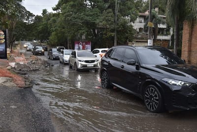 Seis autos atascados en agua, calle deteriorada con escombros y área de construcción a la izquierda, día nublado.