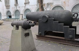 Dos cañones de bronce expuestos al aire libre, uno pequeño en pedestal con placa y otro grande en base metálica.