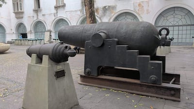 Dos cañones de bronce expuestos al aire libre, uno pequeño en pedestal con placa y otro grande en base metálica.
