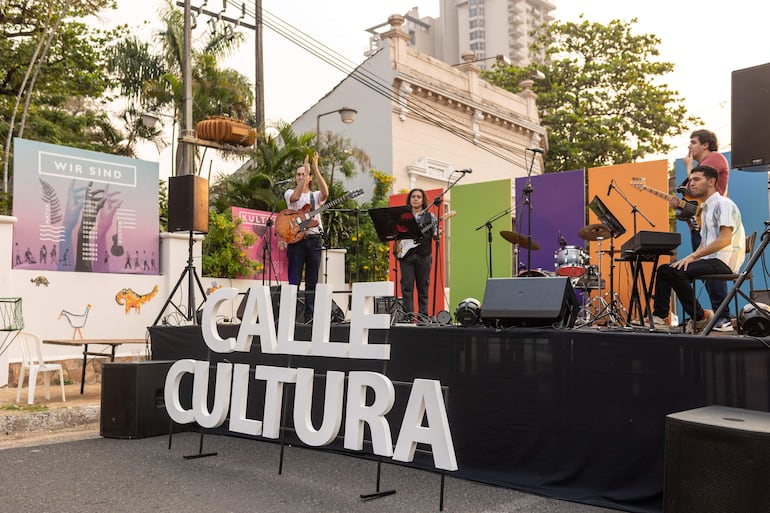 Cuatro músicos en escena: hombre aplaudiendo con guitarra eléctrica, mujer con guitarra, hombre con bajo y otro sentado con teclado.